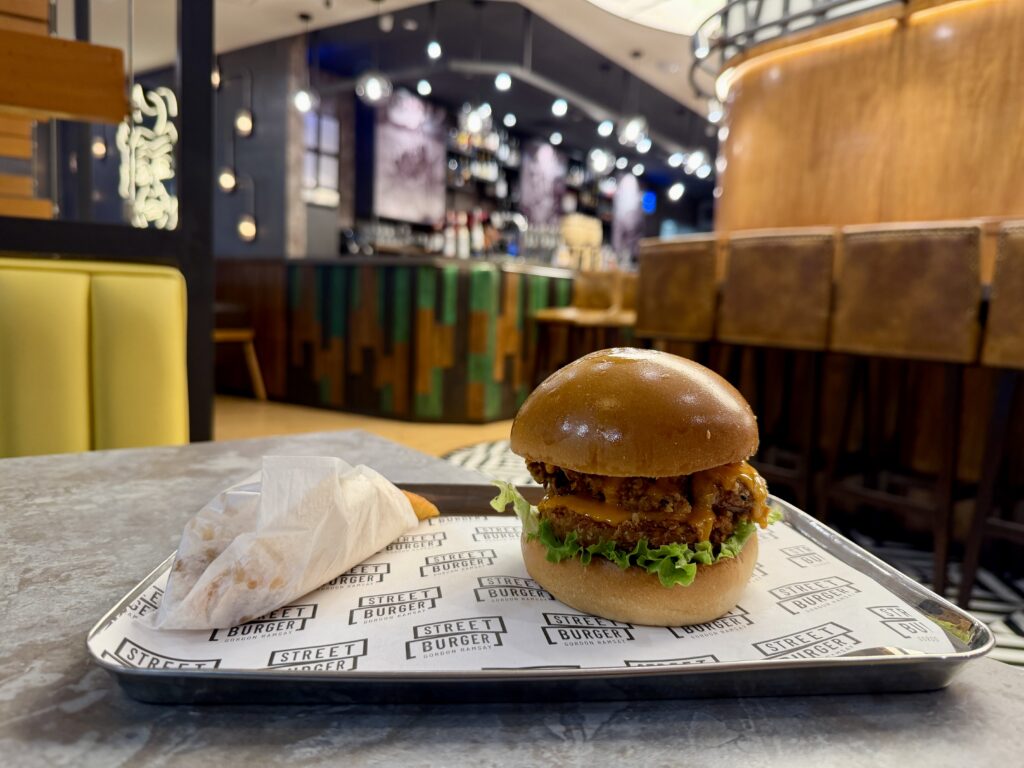 The image shows a close-up of a tray on a table in a restaurant. On the tray, there is a burger with a glossy bun, lettuce, and a patty with sauce. Next to the burger is a wrapped item, possibly fries. The background features a modern restaurant interior with a bar, stools, and decorative lighting.