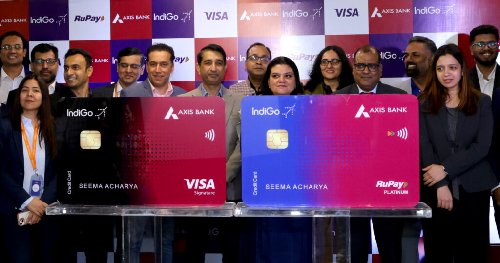 A group of people is standing in front of a backdrop featuring logos of Axis Bank, IndiGo, RuPay, and Visa. They are posing with two large credit card replicas. The cards display the names "Axis Bank" and "IndiGo," with one card labeled "Visa Signature" and the other "RuPay Platinum." The name "Seema Acharya" is printed on both cards.