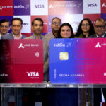 A group of people is standing in front of a backdrop featuring logos of Axis Bank, IndiGo, RuPay, and Visa. They are posing with two large credit card replicas. The cards display the names "Axis Bank" and "IndiGo," with one card labeled "Visa Signature" and the other "RuPay Platinum." The name "Seema Acharya" is printed on both cards.