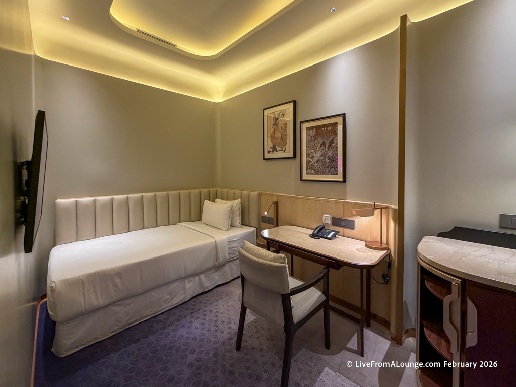 A small, modern hotel room with a single bed against the wall, featuring a padded headboard. There is a desk with a chair, a telephone, and a notepad. Two framed artworks hang above the desk. The room has soft, ambient lighting from the ceiling, and a wall-mounted TV is visible on the left. The decor is minimalist with neutral tones.