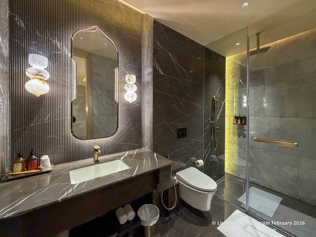 The image shows a modern, luxurious bathroom with dark marble walls and flooring. There is a large, uniquely shaped mirror above a marble countertop with a sink. On the countertop, there are toiletries and a towel. The bathroom features a toilet and a glass-enclosed shower area with a rainfall showerhead and a shelf holding bottles. The lighting is warm, creating an elegant ambiance.