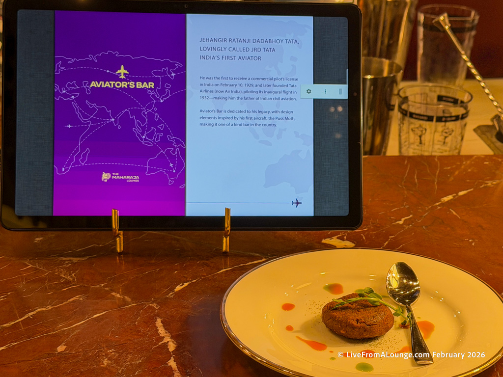 The image shows a tablet on a stand displaying information about "Aviator's Bar" and Jehangir Ratanji Dadabhoy Tata, India's first aviator. The screen has a purple background with a map and text. In front of the tablet, there is a white plate with a round food item garnished with greens and colorful sauce, accompanied by a spoon. The setting appears to be a lounge or bar area.