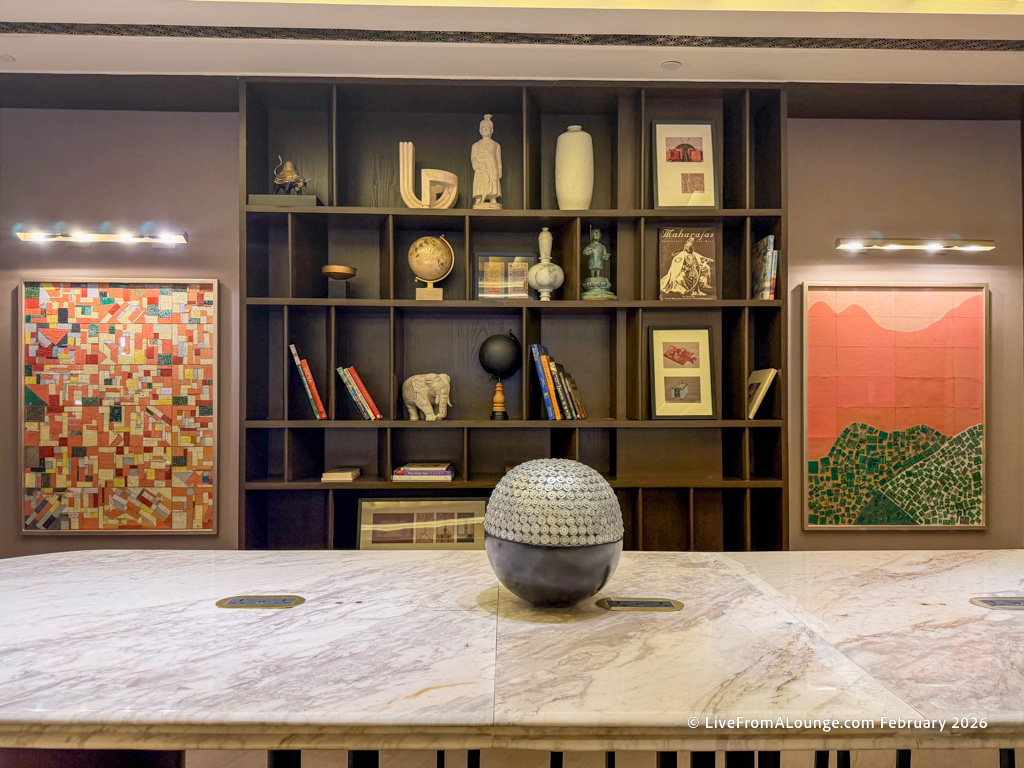 The image shows a modern interior with a large marble table in the foreground. Behind the table is a dark wooden bookshelf filled with various decorative items, including books, framed pictures, a globe, sculptures, and vases. On either side of the bookshelf are two colorful abstract paintings. The lighting is warm, with wall-mounted lights illuminating the artwork.