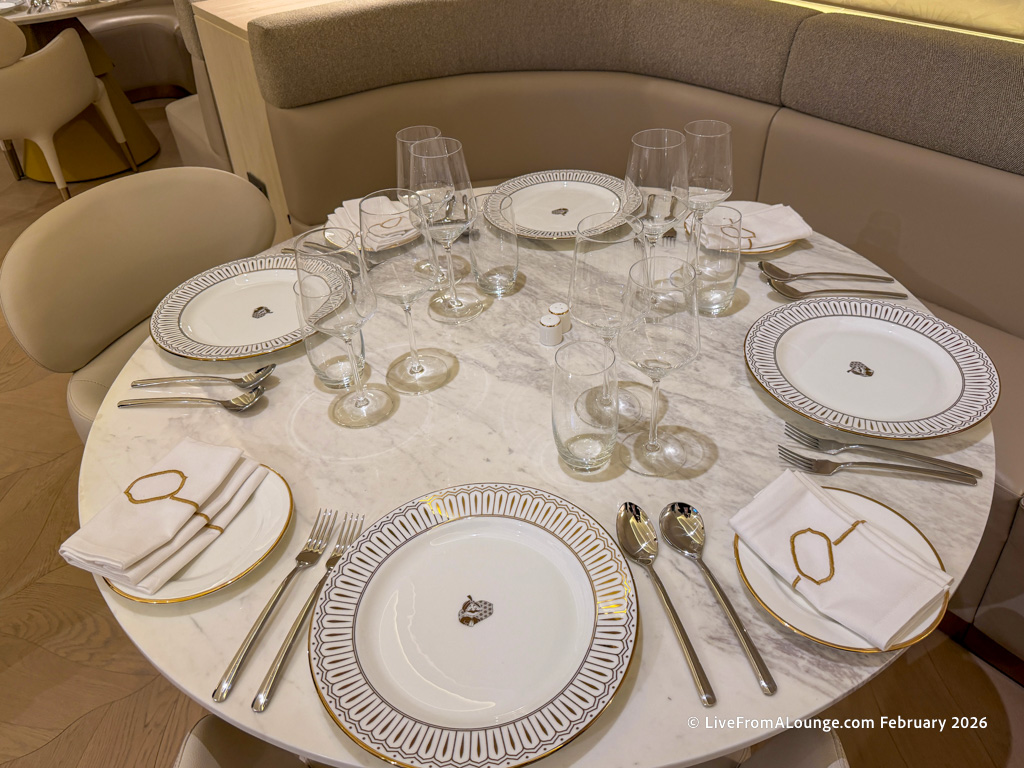 A round marble dining table is elegantly set for four people. Each place setting includes a white plate with a decorative border, a neatly folded napkin with a gold ring, and silverware consisting of a fork, knife, and spoon. There are multiple wine glasses and water glasses arranged in the center of the table. The seating consists of cushioned chairs and a curved bench. The overall setting is sophisticated and ready for a formal meal.