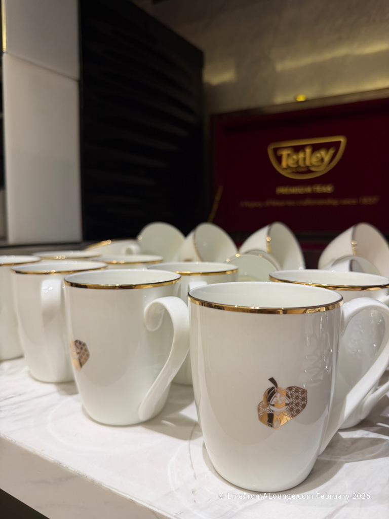 The image shows a collection of white ceramic mugs with gold rims and a decorative gold pattern on the side. They are arranged on a white surface. In the background, there is a red sign with the Tetley logo.