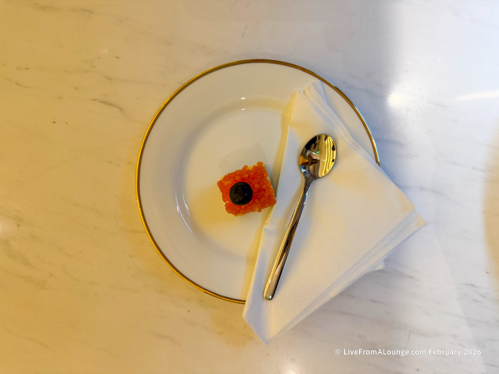 A small dessert topped with a blueberry is placed on a white plate with a gold rim. A folded white napkin and a small spoon are also on the plate, which is set on a light-colored marble surface.