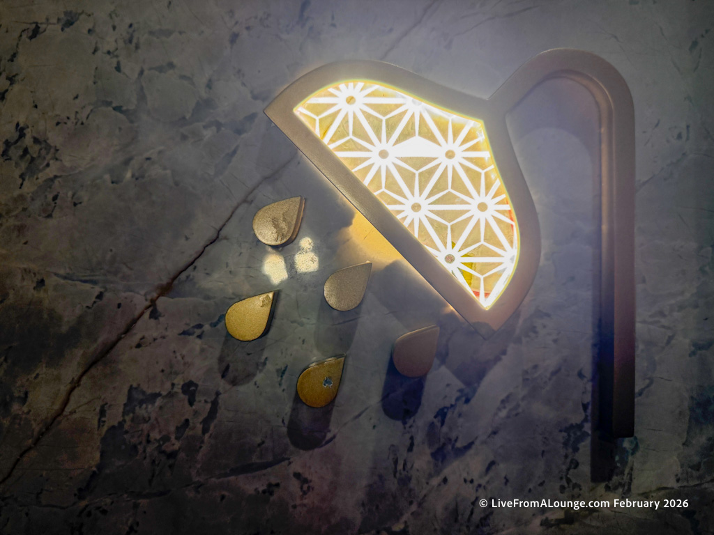 The image shows a decorative wall light shaped like a showerhead, with a geometric pattern that is illuminated. Below the showerhead, there are several teardrop-shaped elements resembling water droplets. The background is a textured, dark surface.