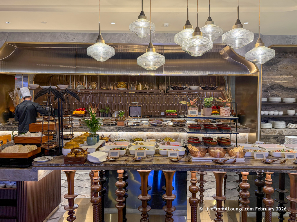 The image shows a buffet setup in a restaurant. There are various dishes displayed in bowls and trays on a marble counter. A chef in a black uniform and white hat is working on the left side. Above the counter, there are several decorative pendant lights. The background features a metallic kitchen hood and shelves with additional food items and decorative elements.