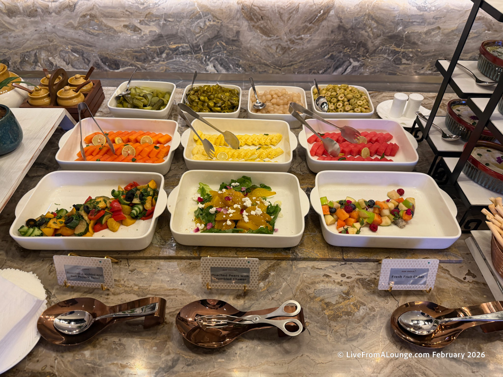 A buffet table with various dishes in white serving trays. The dishes include sliced carrots with lemon, pickles, olives, lychees, watermelon, and pineapple. There are also mixed vegetable salads, a salad with oranges and cheese, and a fresh fruit salad with berries and melon. Copper serving utensils are placed in front of the trays.