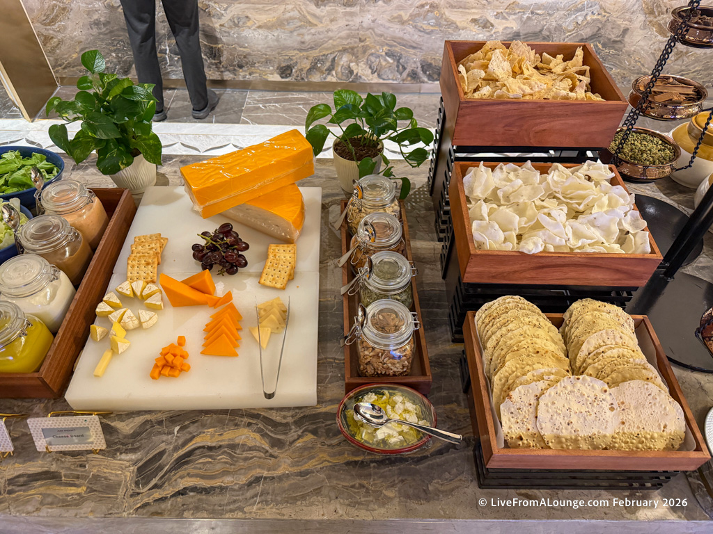 A buffet table featuring a variety of cheeses, crackers, and accompaniments. There are different types of sliced and cubed cheeses on a white board, along with crackers and grapes. To the right, there are wooden trays filled with crispy snacks and round flatbreads. Glass jars containing various condiments and nuts are also present. Green plants are used as decoration on the table.
