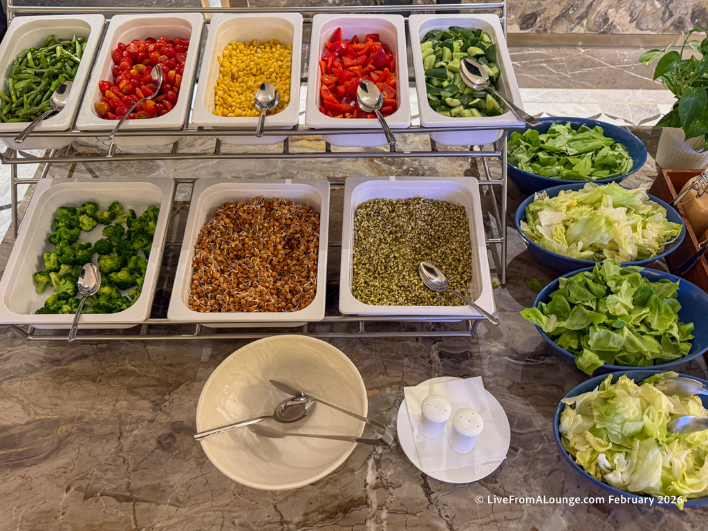 The image shows a salad bar with various fresh ingredients. There are trays containing green beans, cherry tomatoes, corn, red bell peppers, cucumbers, broccoli, and sprouts. To the side, there are bowls filled with different types of lettuce. A bowl with a serving spoon and a small plate with salt and pepper shakers are also visible on the counter.