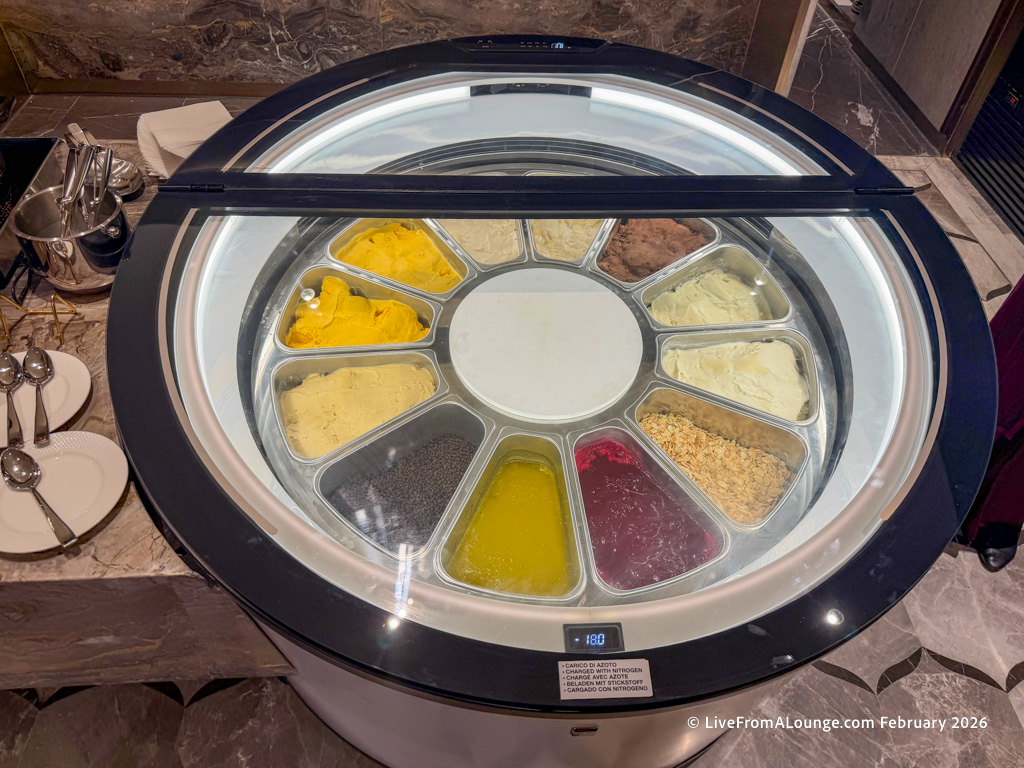 The image shows a circular ice cream display freezer with multiple compartments, each containing different flavors of ice cream and toppings. The freezer is set into a countertop, and there are spoons and small plates placed nearby for serving.