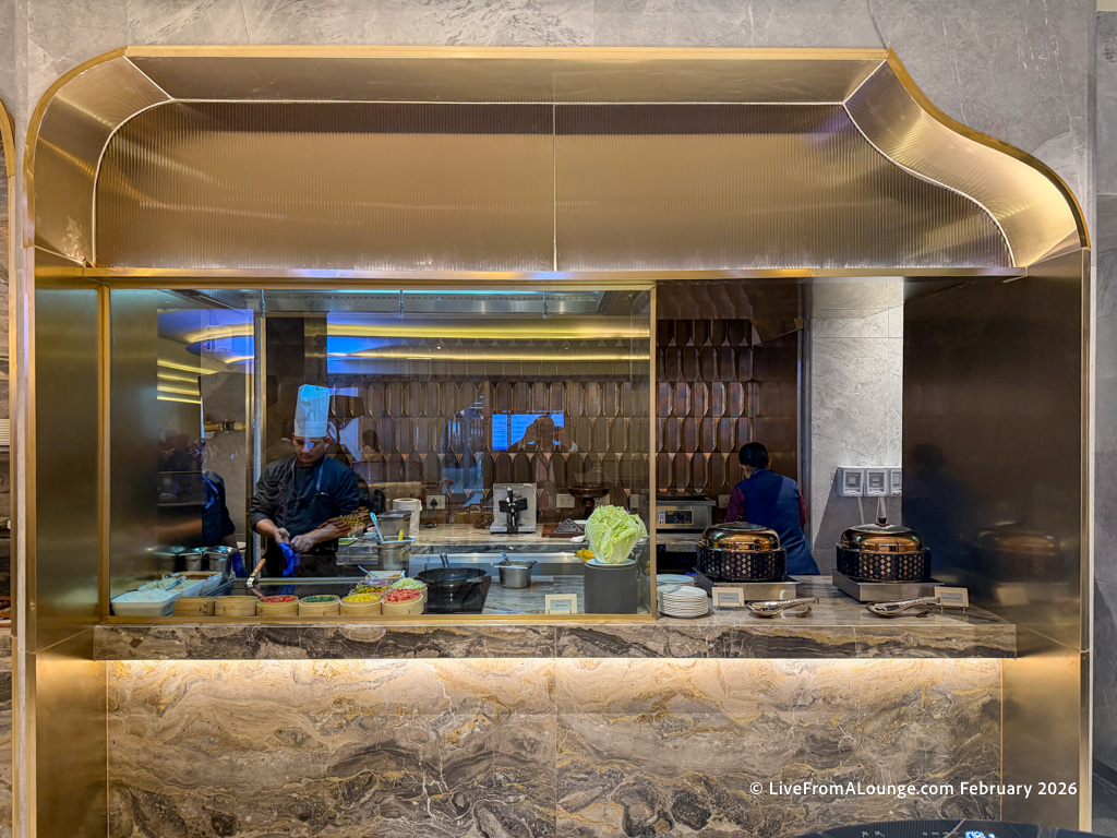 The image shows a modern kitchen with a chef wearing a white hat and dark uniform preparing food. The kitchen has a marble countertop with various ingredients and cooking utensils. There are two covered dishes on the right side of the counter. The background features a decorative wall and a reflection of the dining area.