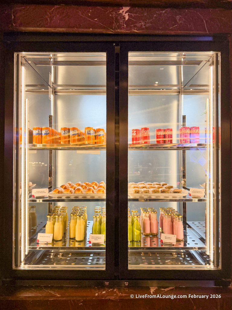 The image shows a refrigerated display case with two glass doors. Inside, there are several shelves containing various beverages and snacks. The top shelf has cans of Fanta and Coca-Cola. The middle shelf holds trays of pastries and small desserts. The bottom shelf contains bottles of different colored drinks, including yellow, green, and pink. Each item is neatly arranged, and there are small labels in front of some items.