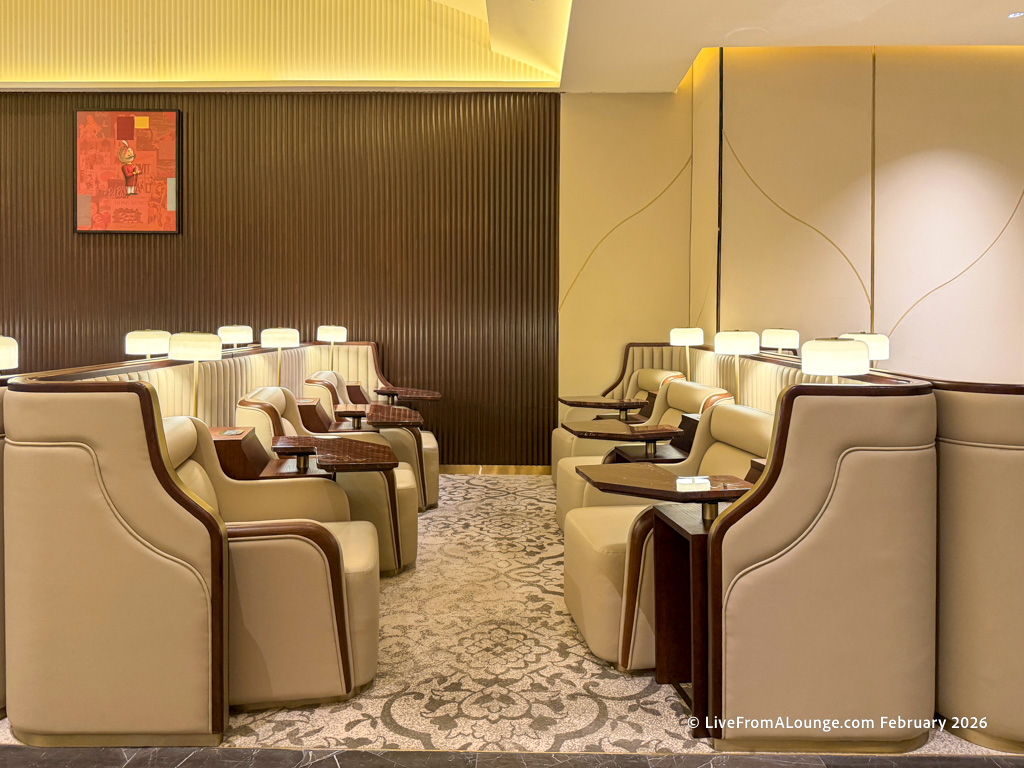 The image shows a modern, well-lit lounge area with rows of beige leather chairs featuring wooden armrests and small side tables. The floor is covered with a patterned carpet, and there are wall-mounted lamps providing soft lighting. The walls are decorated with a combination of dark wood paneling and light-colored panels, and there is a piece of abstract art hanging on one wall.