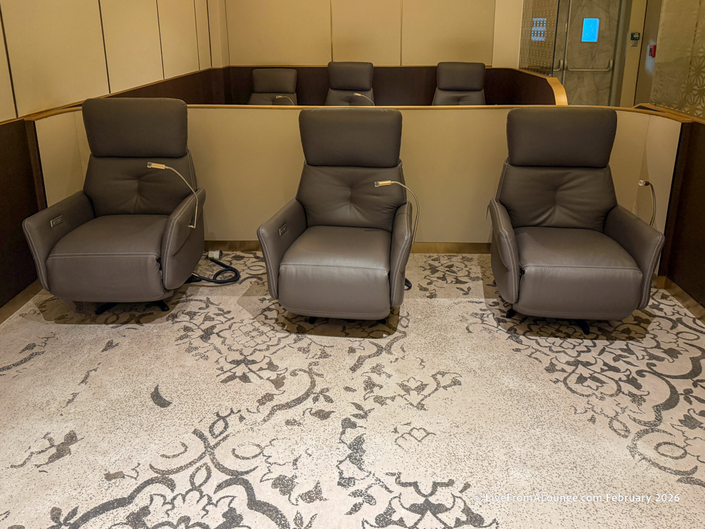 The image shows a room with three gray reclining chairs arranged in a row. Each chair has an attached reading light. The floor is covered with a patterned carpet, and there is a partition wall behind the chairs. The setting appears to be a lounge or waiting area.