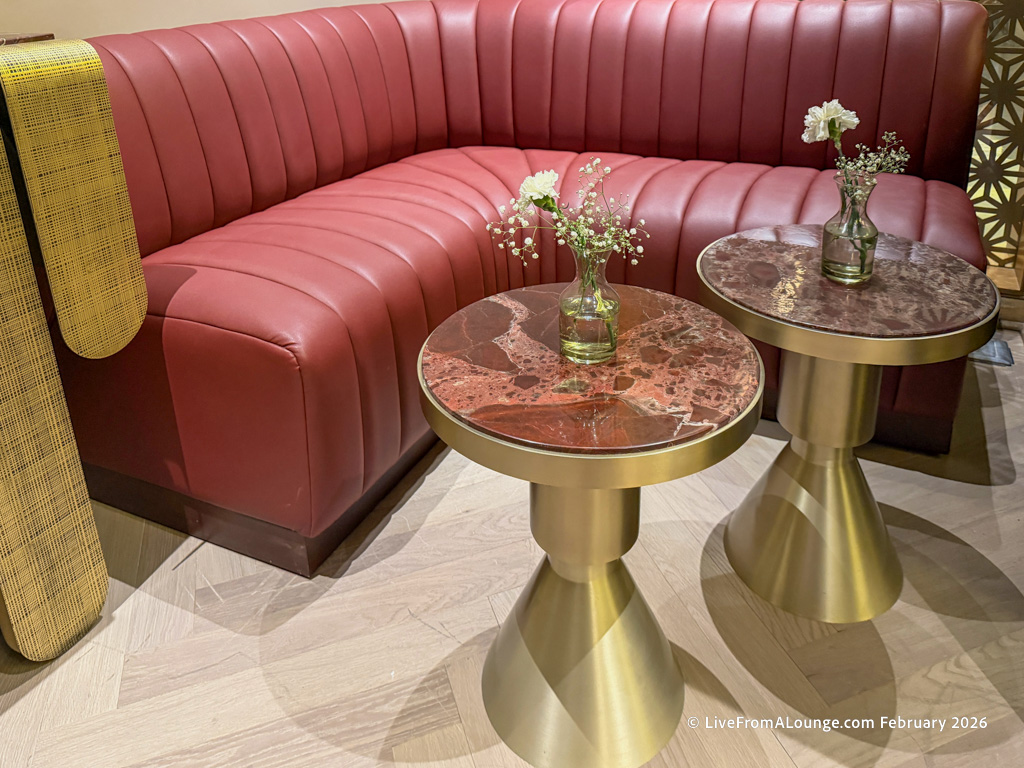A stylish seating area featuring a red, tufted leather sofa and two round, marble-topped tables with gold bases. Each table has a small glass vase with white flowers. The floor is light wood, and a patterned yellow fabric is draped over the sofa's arm.