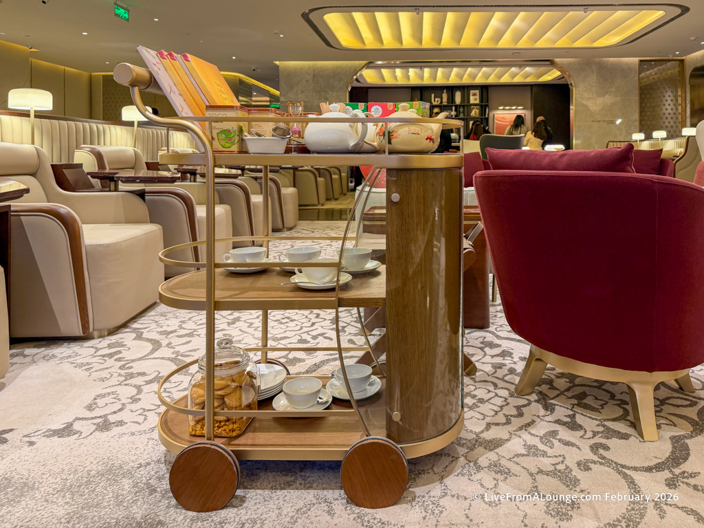 A luxurious lounge setting featuring a tea cart with various items. The cart has a wooden and metal design, holding teapots, cups, and a jar of cookies. The background shows plush seating with beige and red chairs, soft lighting, and a patterned carpet. The atmosphere is elegant and inviting.