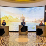 The image shows three people standing behind separate podiums in a welcoming gesture, with their hands together in front of a large, curved digital screen. The screen displays a scenic background of a lake with lotus flowers, surrounded by mountains and architectural structures. The floor has a circular, patterned design, and there are lamps on the podiums. The setting appears to be a reception area, possibly in a lounge or hotel.