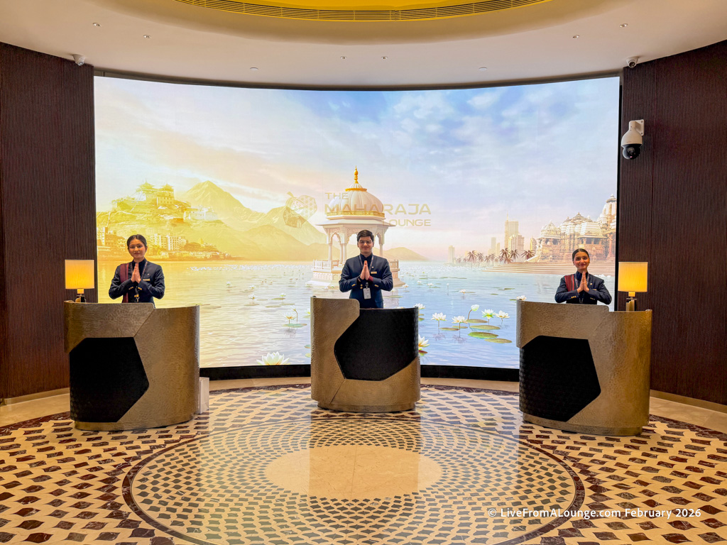 Three people in formal attire stand behind separate podiums, each with their hands in a traditional greeting gesture. They are positioned in front of a large, curved screen displaying an artistic backdrop of a serene water scene with mountains and architectural structures. The floor features a circular mosaic pattern, and there are lamps on the podiums. The setting appears to be a welcoming area, possibly in a lounge or reception space.