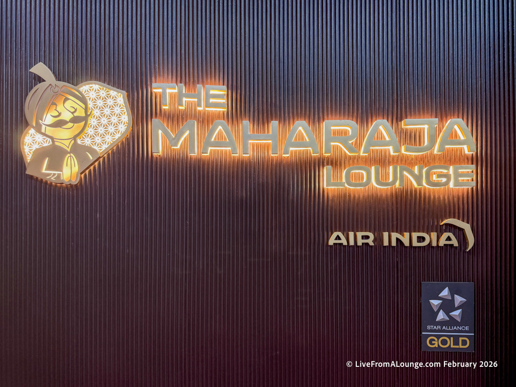 The image shows a sign for "The Maharaja Lounge" associated with Air India. The sign features a stylized illustration of a character with folded hands, resembling a traditional Indian greeting. The text "Air India" and the Star Alliance Gold logo are also visible. The background is a textured, dark surface.