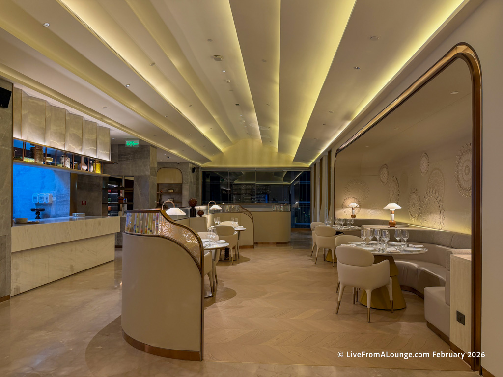 The image shows an elegant and modern restaurant interior. The space features a high ceiling with soft, ambient lighting. There are several round tables set with white tablecloths, glassware, and table lamps. The walls have decorative patterns, and the floor is made of polished wood. The overall atmosphere is sophisticated and inviting.