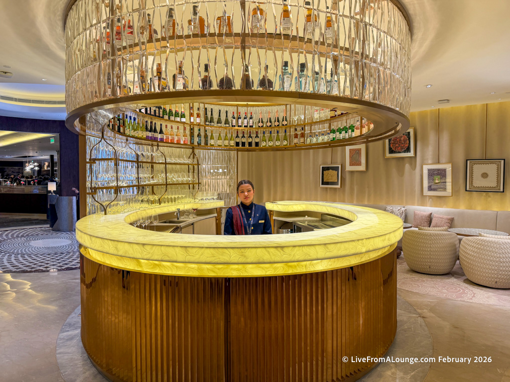 The image shows a luxurious circular bar with a glowing yellow countertop and a decorative glass rack above, filled with various bottles. A person in a uniform stands behind the bar. The surrounding area features elegant seating with cushioned chairs and framed artwork on the walls, creating a sophisticated ambiance.