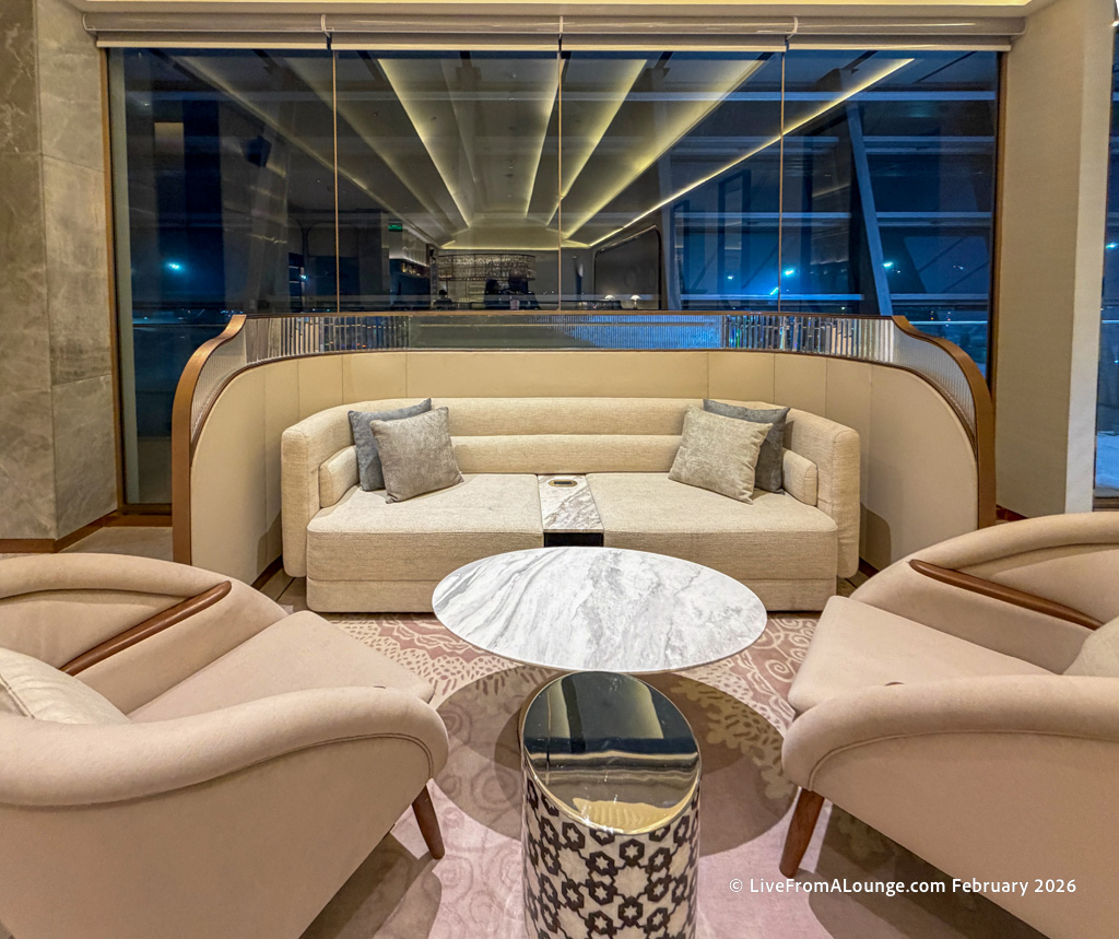 A modern lounge area featuring a curved beige sofa with two gray cushions, two matching armchairs, and a round marble coffee table. The room has a large window with a view of the outside, and the ceiling has recessed lighting. The decor is elegant and contemporary.