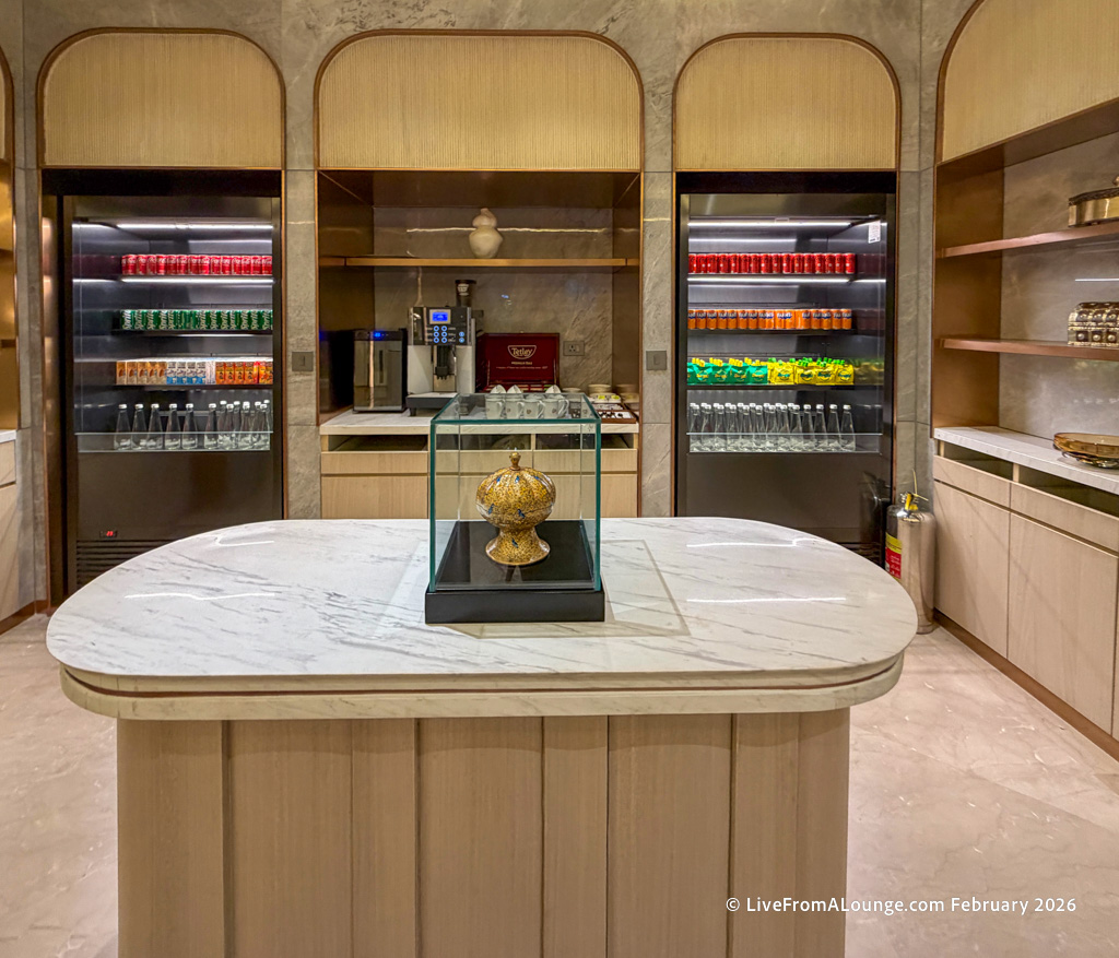 The image shows a modern kitchen or pantry area with a marble countertop in the foreground. On the countertop, there is a decorative object inside a glass case. In the background, there are two refrigerators filled with various beverages, including cans and bottles. Between the refrigerators, there is a coffee machine and a shelf with cups. The walls and cabinetry have a light wood finish, and there are additional shelves with items on the right side.