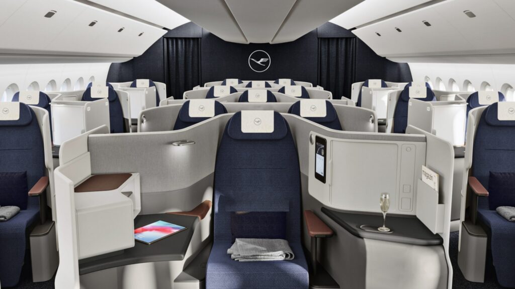 This image shows the interior of an airplane's business class cabin. The seats are arranged in a spacious layout with privacy partitions. Each seat has a personal entertainment screen, a small table with a tablet, and a glass of champagne. The seats are upholstered in dark blue fabric with light gray and brown accents. The cabin has a modern and luxurious design, with overhead storage compartments and windows along the sides.