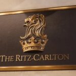The image shows a plaque with a gold border featuring a gold lion's head above a crown. Below the emblem, the text reads "The Ritz-Carlton."
