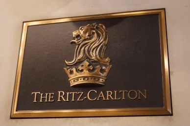 The image shows a plaque with a gold border featuring a gold lion's head above a crown. Below the emblem, the text reads "The Ritz-Carlton."