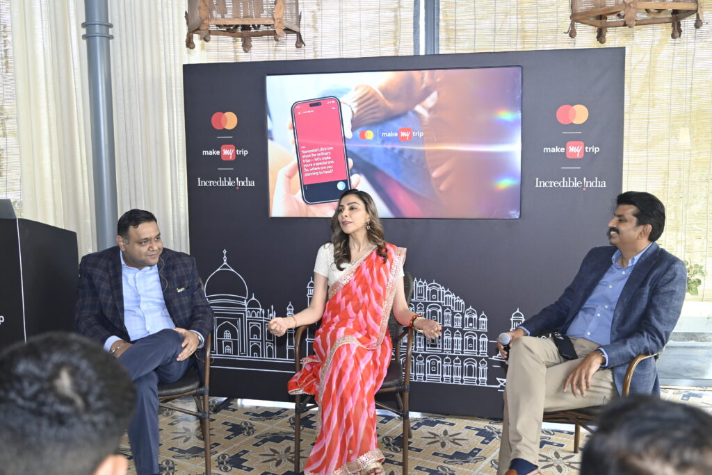 Three people are seated in chairs, engaged in a discussion. The person in the center is wearing a red and white saree, while the two others are in suits. Behind them is a screen displaying a smartphone with the MakeMyTrip logo and text. The backdrop features the logos of MakeMyTrip and Incredible India, along with illustrations of Indian architecture.