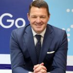 A man in a blue suit and tie is sitting with his hands clasped. He is smiling and positioned in front of a blue and white background with partial text visible.