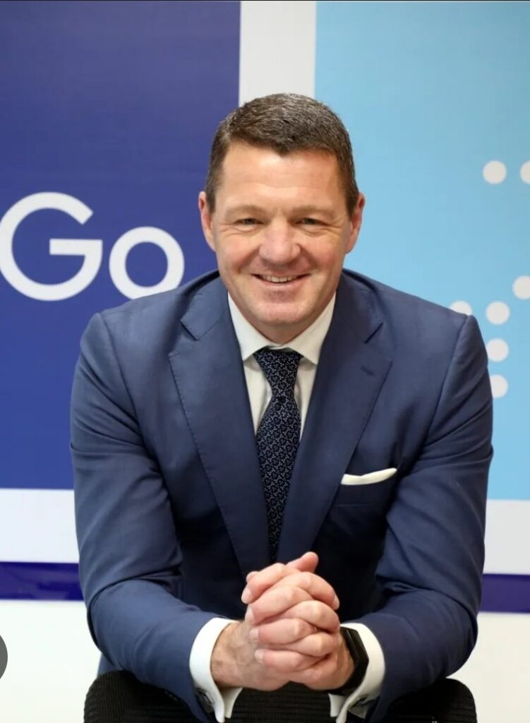 A man in a blue suit and tie is sitting with his hands clasped. He is smiling and positioned in front of a blue and white background with partial text visible.