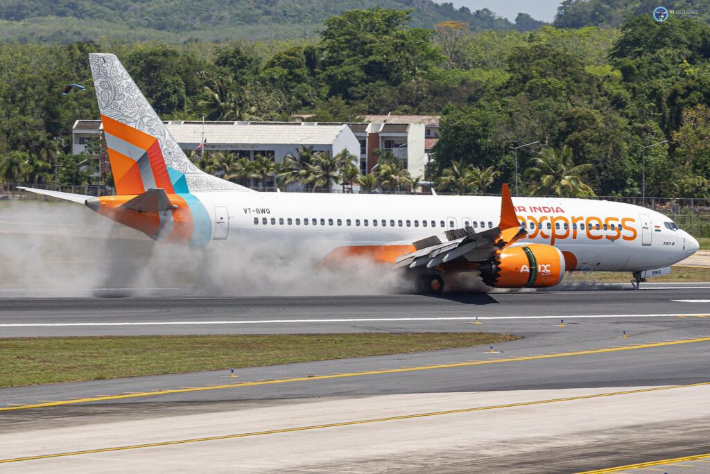 The image shows an Air India Express airplane landing on a runway. The aircraft is a Boeing 737, and its wheels are touching down, creating a cloud of smoke. The plane has a colorful tail design with orange, teal, and white patterns. In the background, there are trees and a building.