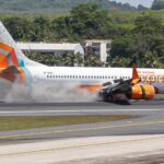The image shows an Air India Express airplane landing on a runway. The aircraft is a Boeing 737, and its wheels are touching down, creating a cloud of smoke. The plane has a colorful tail design with orange, teal, and white patterns. In the background, there are trees and a building.