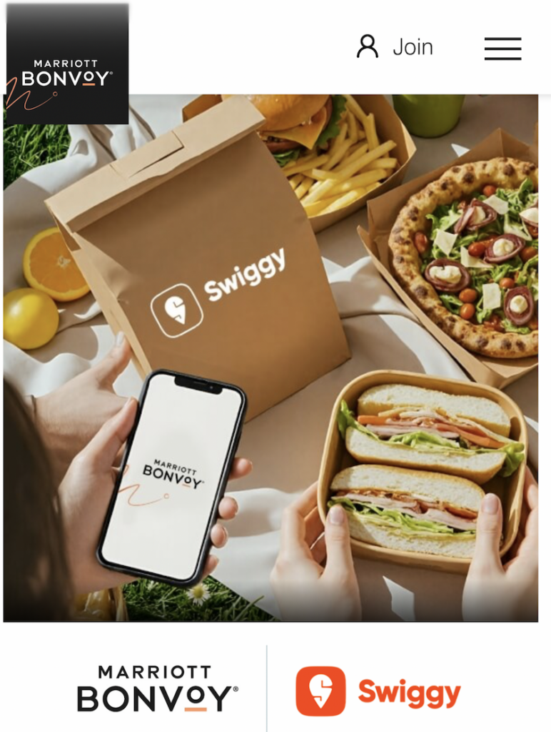The image shows a picnic setup with various food items, including a pizza, a burger with fries, and a sandwich. A person is holding a smartphone displaying the Marriott Bonvoy logo. There is a Swiggy-branded paper bag, indicating a collaboration between Marriott Bonvoy and Swiggy. The background includes fruits like oranges and lemons.