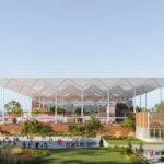The image shows a modern architectural structure with a large, white, tent-like roof supported by tall pillars. Below the roof, there are green spaces and trees, with people walking around a plaza area. The foreground features landscaped gardens and a small building with a circular logo on its facade. The sky is clear and blue, suggesting a sunny day.