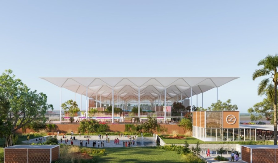 The image shows a modern architectural structure with a large, white, tent-like roof supported by tall pillars. Below the roof, there are green spaces and trees, with people walking around a plaza area. The foreground features landscaped gardens and a small building with a circular logo on its facade. The sky is clear and blue, suggesting a sunny day.